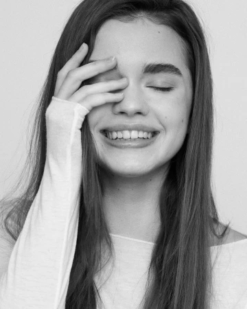 Close-up monochrome portrait of a smiling young woman with long dark hair, holding her hand over her eye against a plain white background.