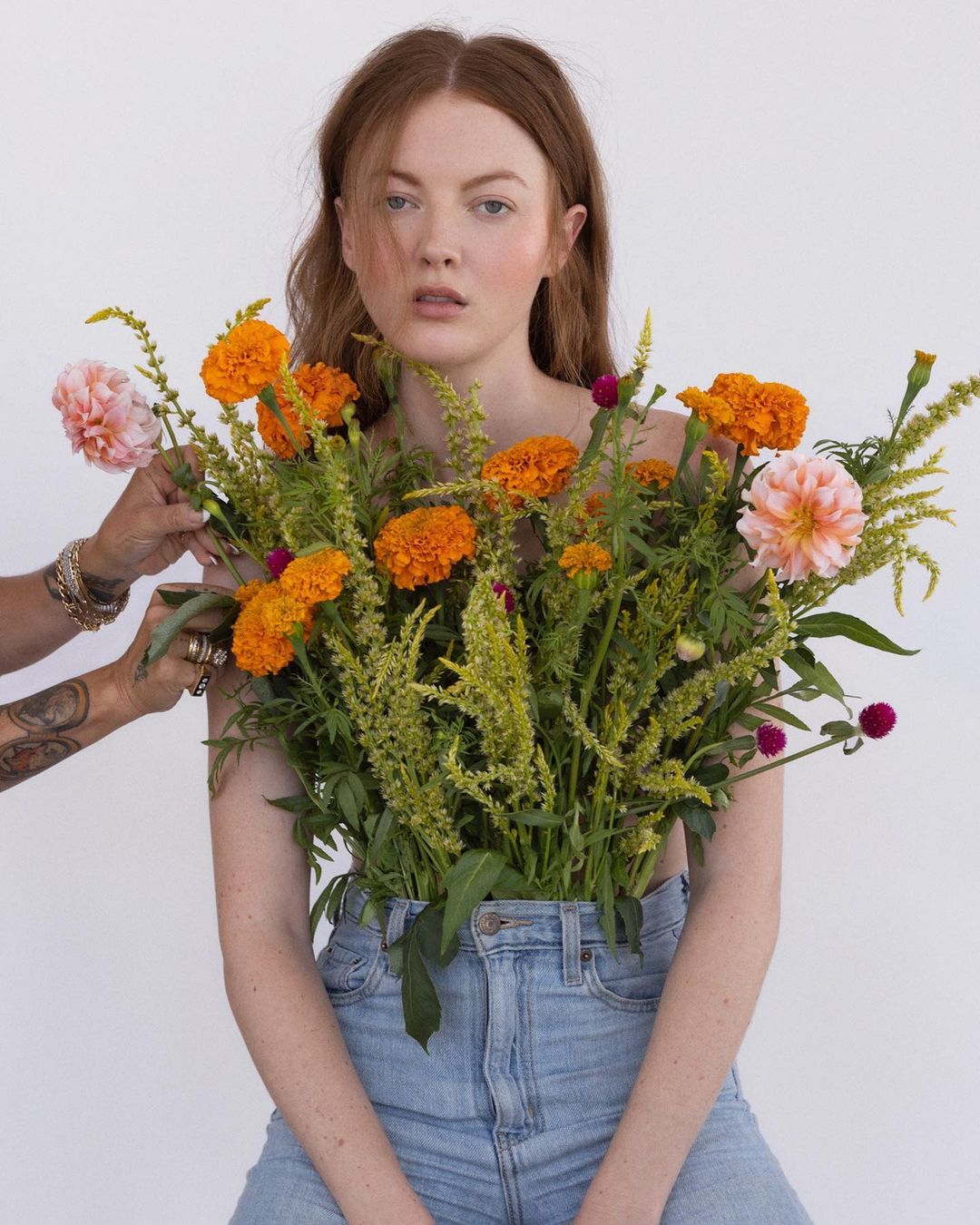 Woman with reddish-brown hair and blue jeans poses with flowers with hand adjusting. Pale background. Medium shot.