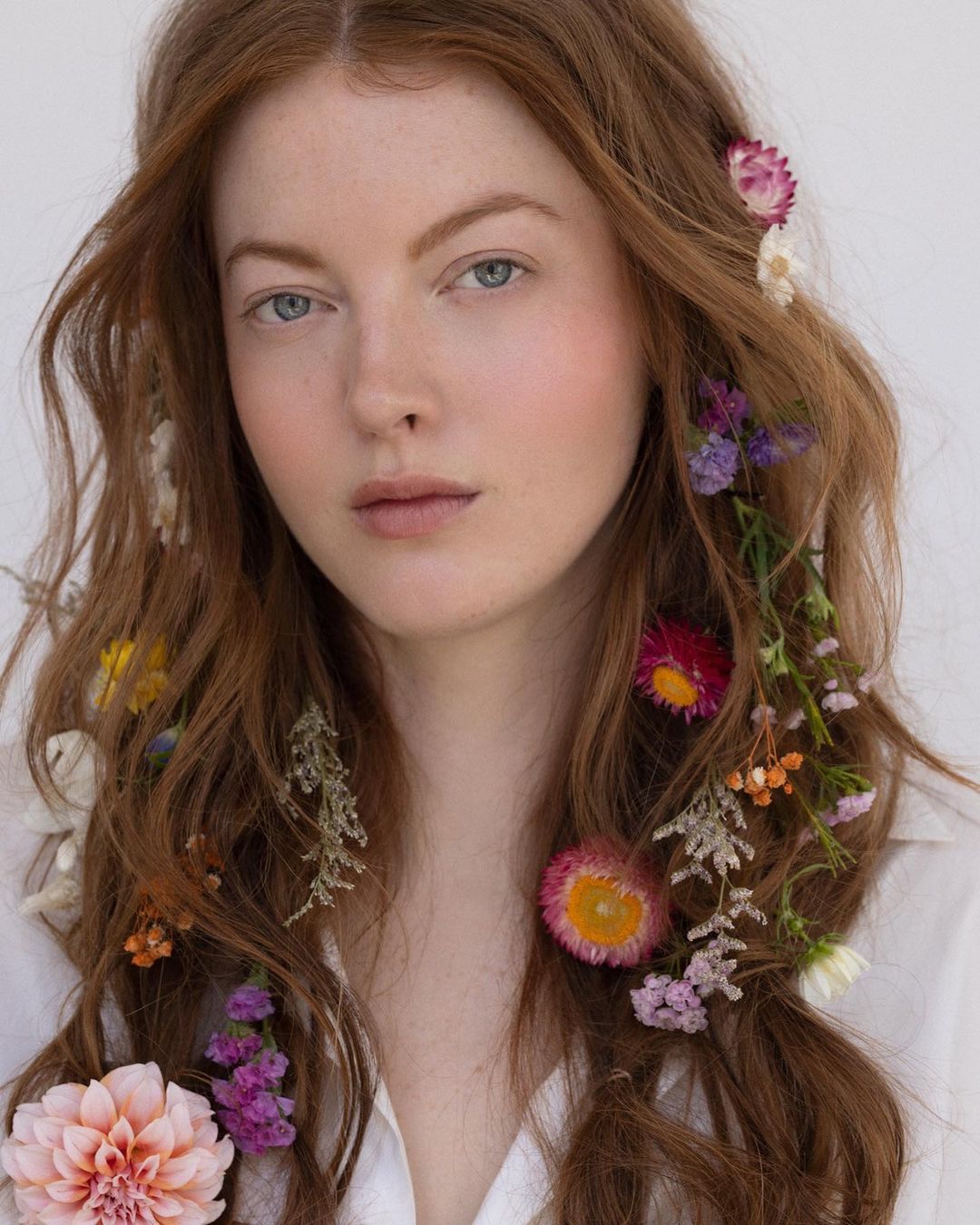 Portrait of a woman with long auburn hair woven with small colorful wildflowers against a plain white background