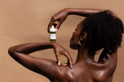 Close-up studio shot of a dark-skinned adult woman posing with a bottle of skincare product, set against a tan backdrop.