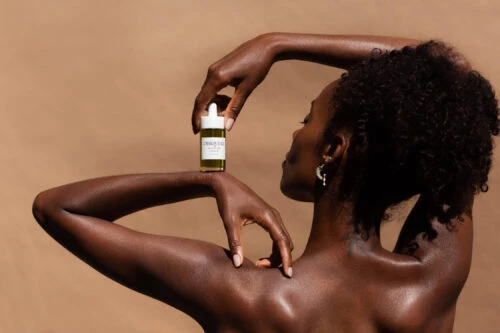 Close-up studio shot of a dark-skinned adult woman posing with a bottle of skincare product, set against a tan backdrop.