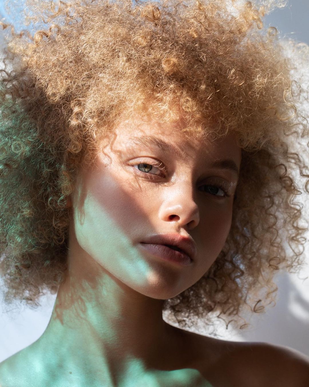 Close-up portrait of a young woman with blonde curly hair, softly lit with a hint of blue light, looking directly at the camera.