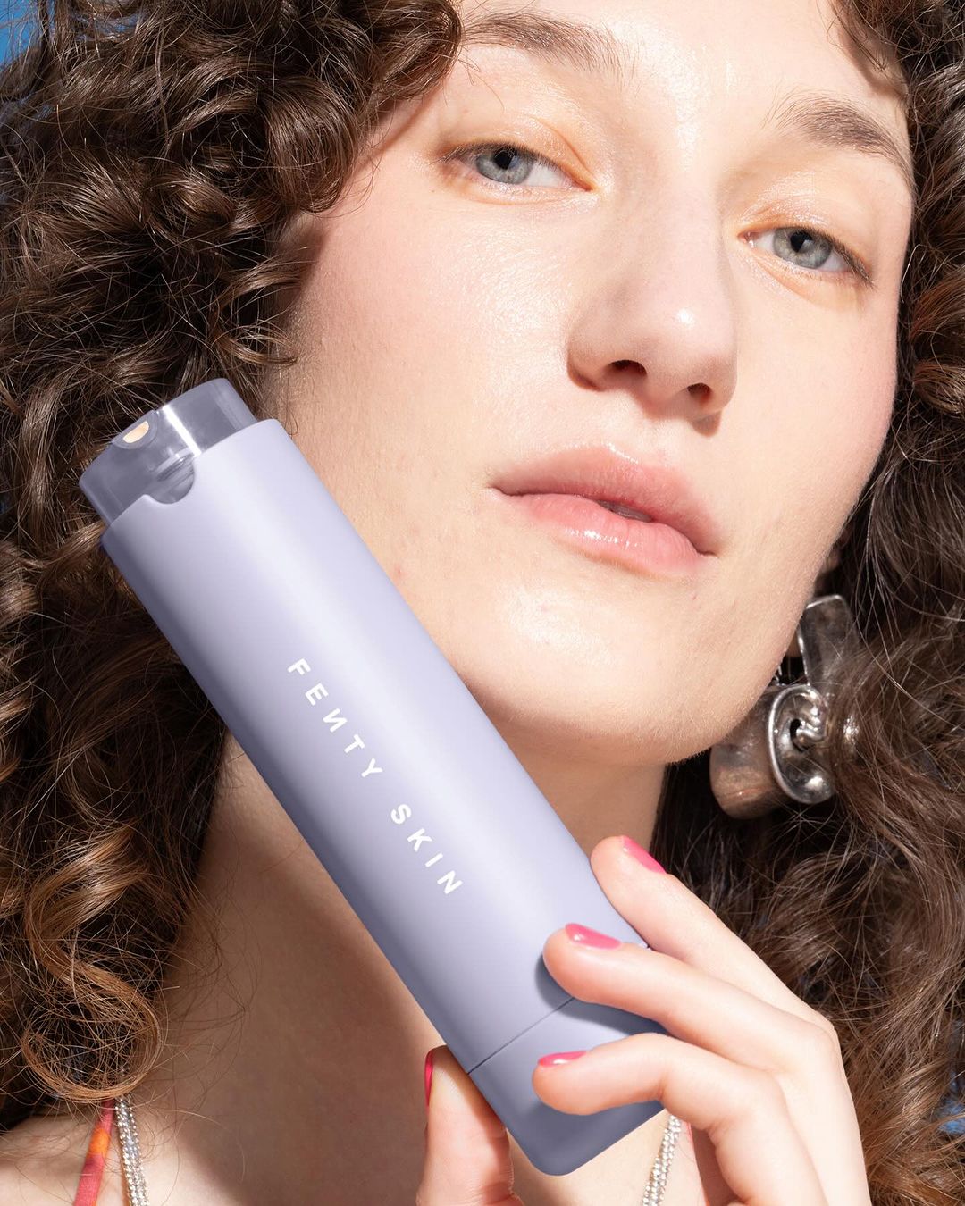 Adult woman holding a purple Fenty Skin skincare product with silver earrings, pink nail polish, and curly hair in portrait shot.