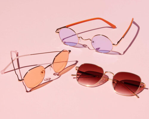 Still life studio shot of three pairs of sunglasses, each with a colorful lens, arranged against a solid light pink background.