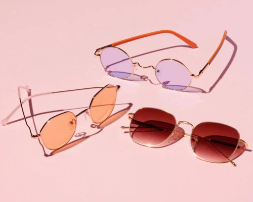 Still life studio shot of three pairs of sunglasses, each with a colorful lens, arranged against a solid light pink background.