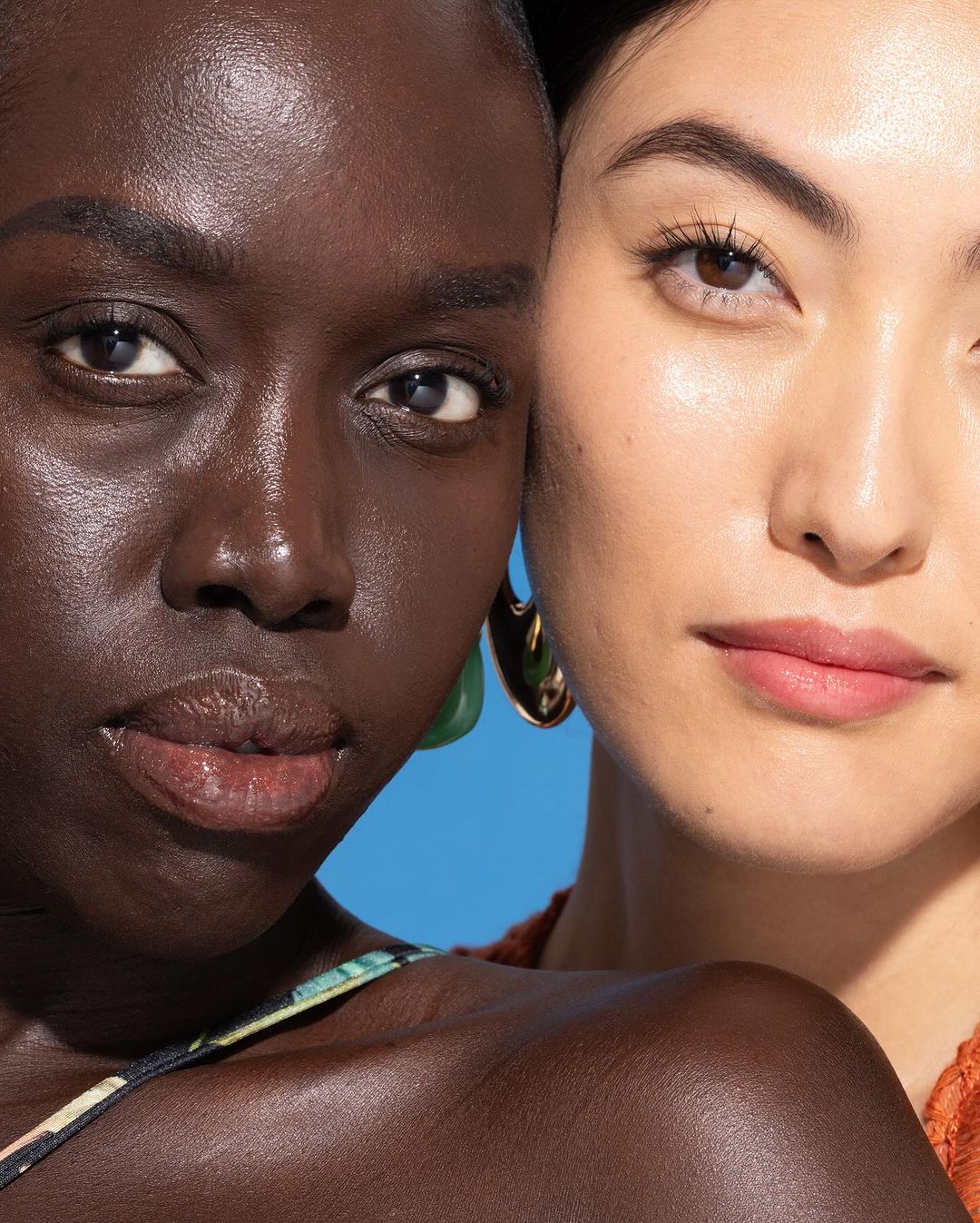 Close-up studio portrait of two adult women with different skin tones looking at camera against light blue background; beauty and fashion.