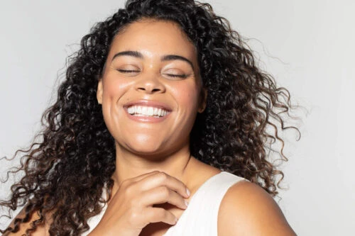 Close-up portrait of a smiling woman with curly dark hair wearing a white top against a neutral background by Angela Marklew.