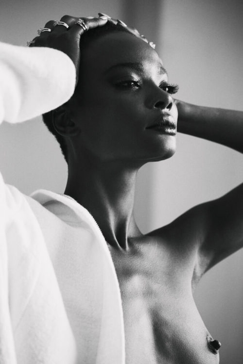Elegant black and white portrait of a topless, dark-skinned woman with her hands on her head, draped with white fabric, studio lit.