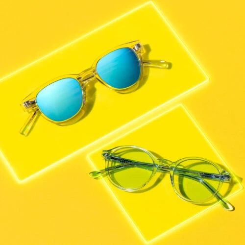 Two pairs of clear-frame sunglasses with blue and green lenses on yellow background, product shot, vibrant lighting, studio photography.