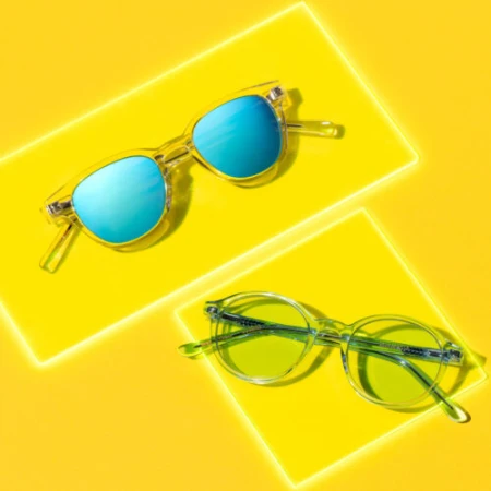 Two pairs of clear-frame sunglasses with blue and green lenses on yellow background, product shot, vibrant lighting, studio photography.