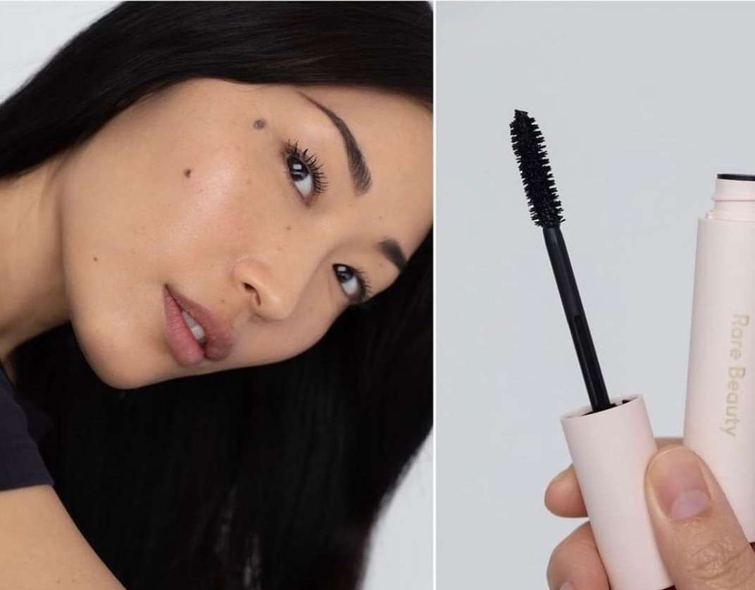 Close-up shot of an adult woman with dark hair wearing mascara, positioned next to a tube of Rare Beauty mascara and applicator wand.