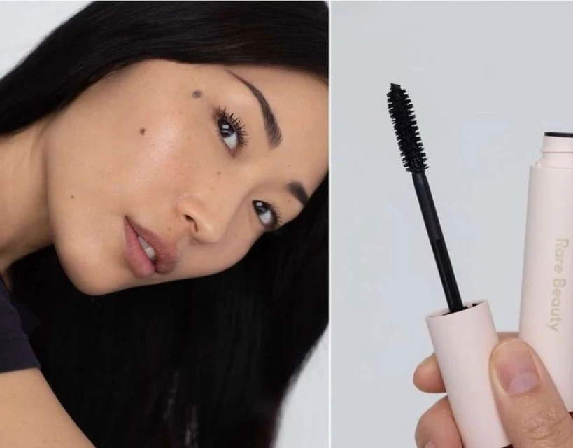 Close-up shot of an adult woman with dark hair wearing mascara, positioned next to a tube of Rare Beauty mascara and applicator wand.