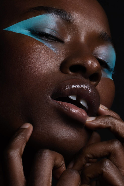 Close-up of a dark-skinned woman with bright blue makeup accentuating her eyes, set against a dark, neutral background.