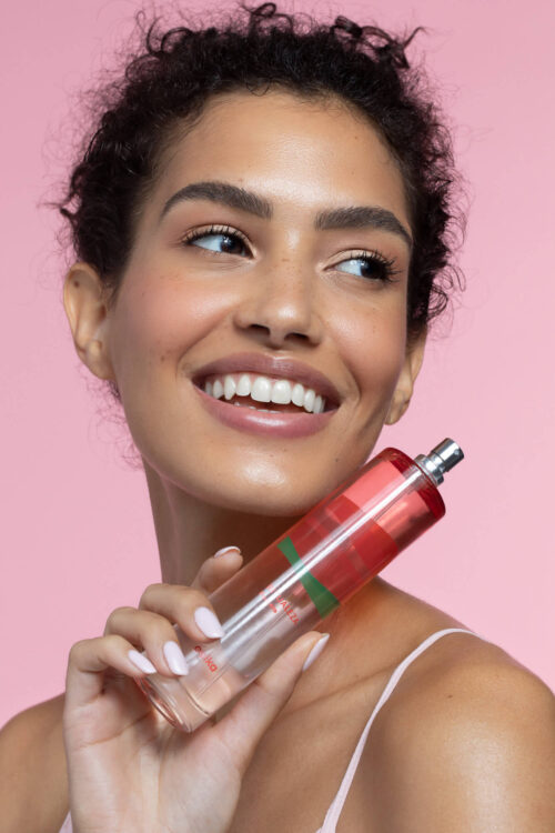 Smiling woman holds a transparent perfume bottle against a light pink background. Beauty, fashion, and cosmetic advertising.