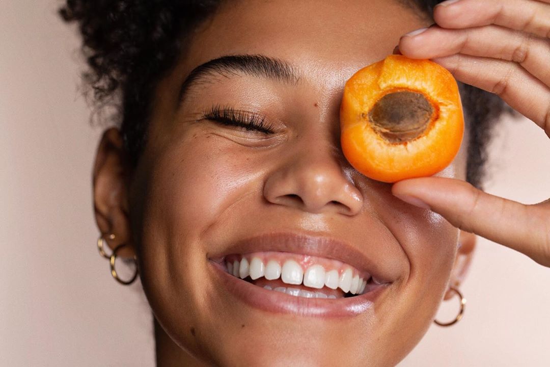 Close-up of a smiling woman with dark curly hair, wearing gold hoop earrings, holding a halved apricot with pit against her eye.