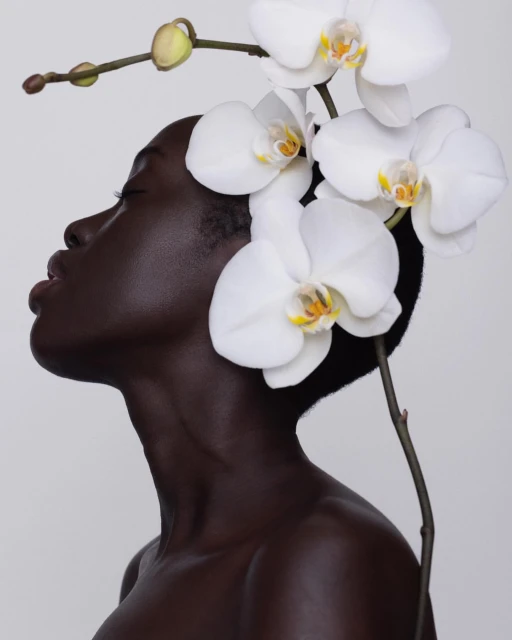 Striking portrait of a Black woman in profile, adorned with white orchids against a soft gray background in a serene fashion beauty shot.