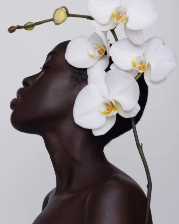 Striking portrait of a Black woman in profile, adorned with white orchids against a soft gray background in a serene fashion beauty shot.