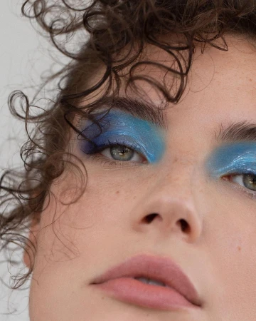 Close-up beauty portrait of a woman with curly brown hair and bold blue metallic eyeshadow by Angela Marklew