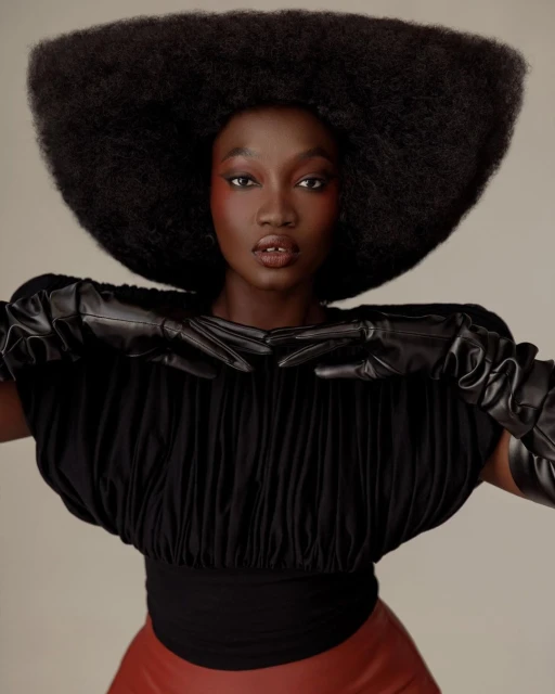 Fashionable African woman poses with large afro, wearing black top and leather gloves, in a studio setting with neutral backdrop.