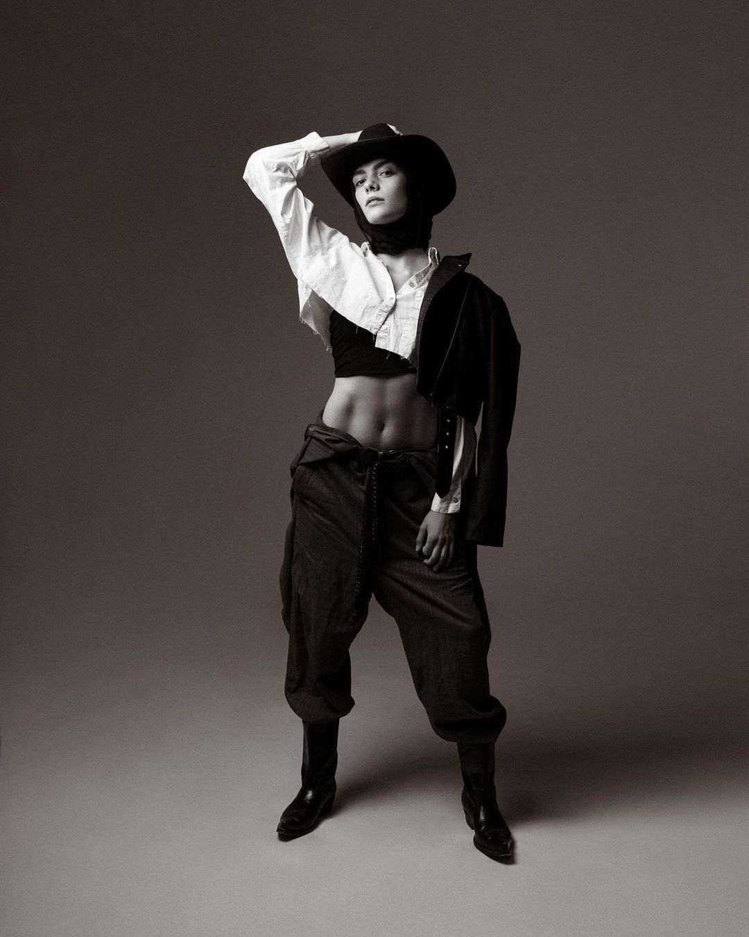 Full-length studio portrait of a person wearing a cowboy hat, crop top, oversized trousers, blazer, and boots, posing with a hand on their head.