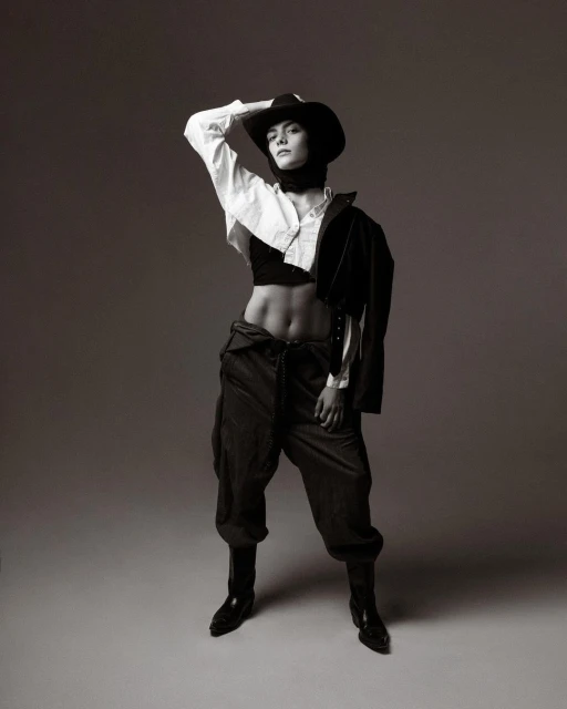 Full-length studio portrait of a person wearing a cowboy hat, crop top, oversized trousers, blazer, and boots, posing with a hand on their head.