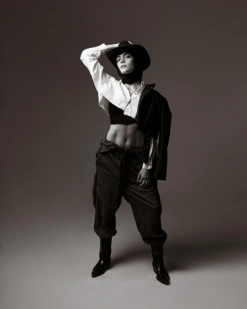Full-length studio portrait of a person wearing a cowboy hat, crop top, oversized trousers, blazer, and boots, posing with a hand on their head.