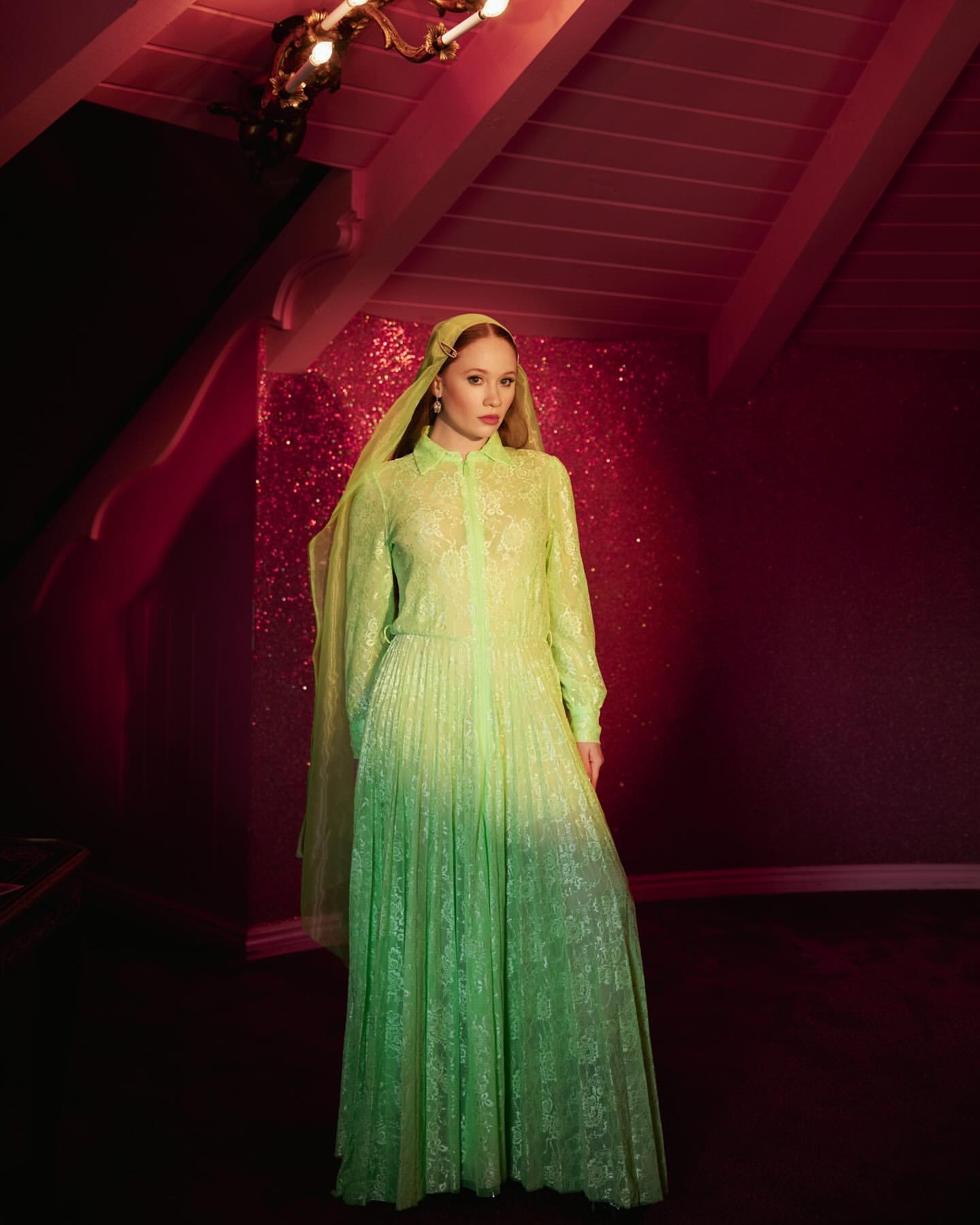 Woman wearing a neon green lace dress and matching headscarf standing in a dimly lit room with a glittery wall and pitched wooden ceiling