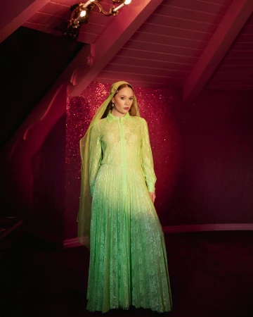Woman wearing a neon green lace dress and matching headscarf standing in a dimly lit room with a glittery wall and pitched wooden ceiling