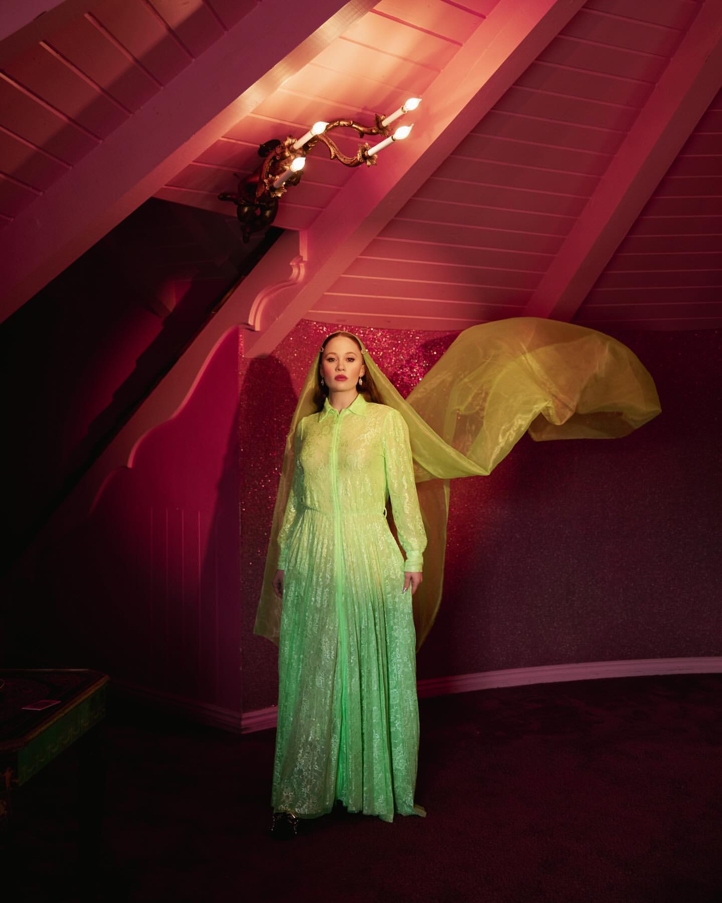 Elegant woman in a lime green gown and sheer veil stands in a pink-toned interior, illuminated by a vintage chandelier, looking at the camera.