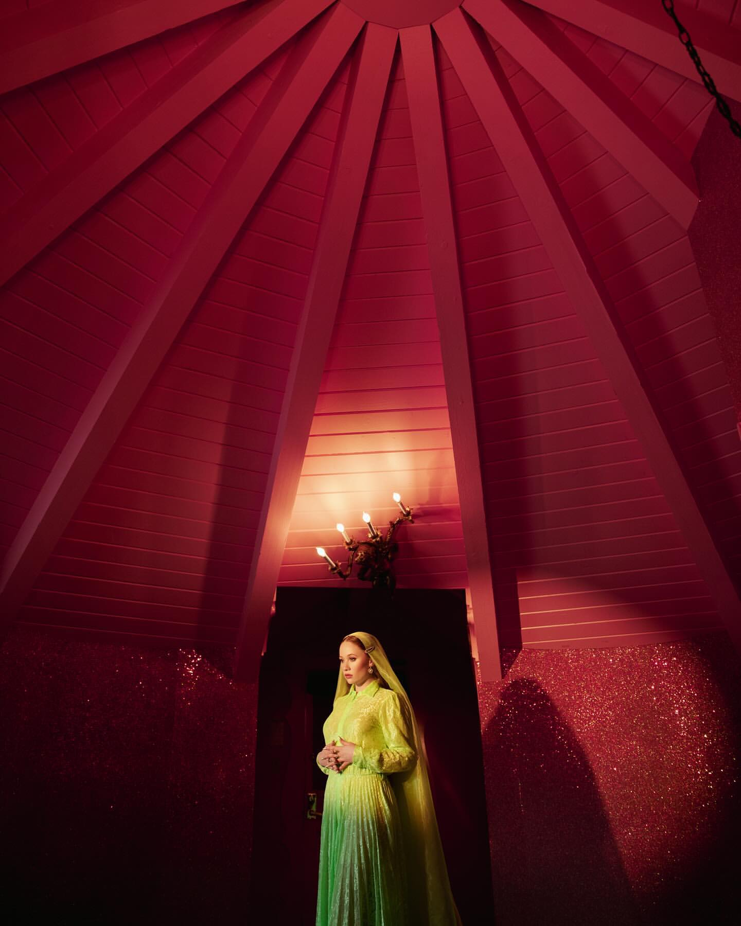 Portrait of a pregnant woman in a green dress standing indoors beneath a unique red ceiling with sconce lighting, in a serene pose.