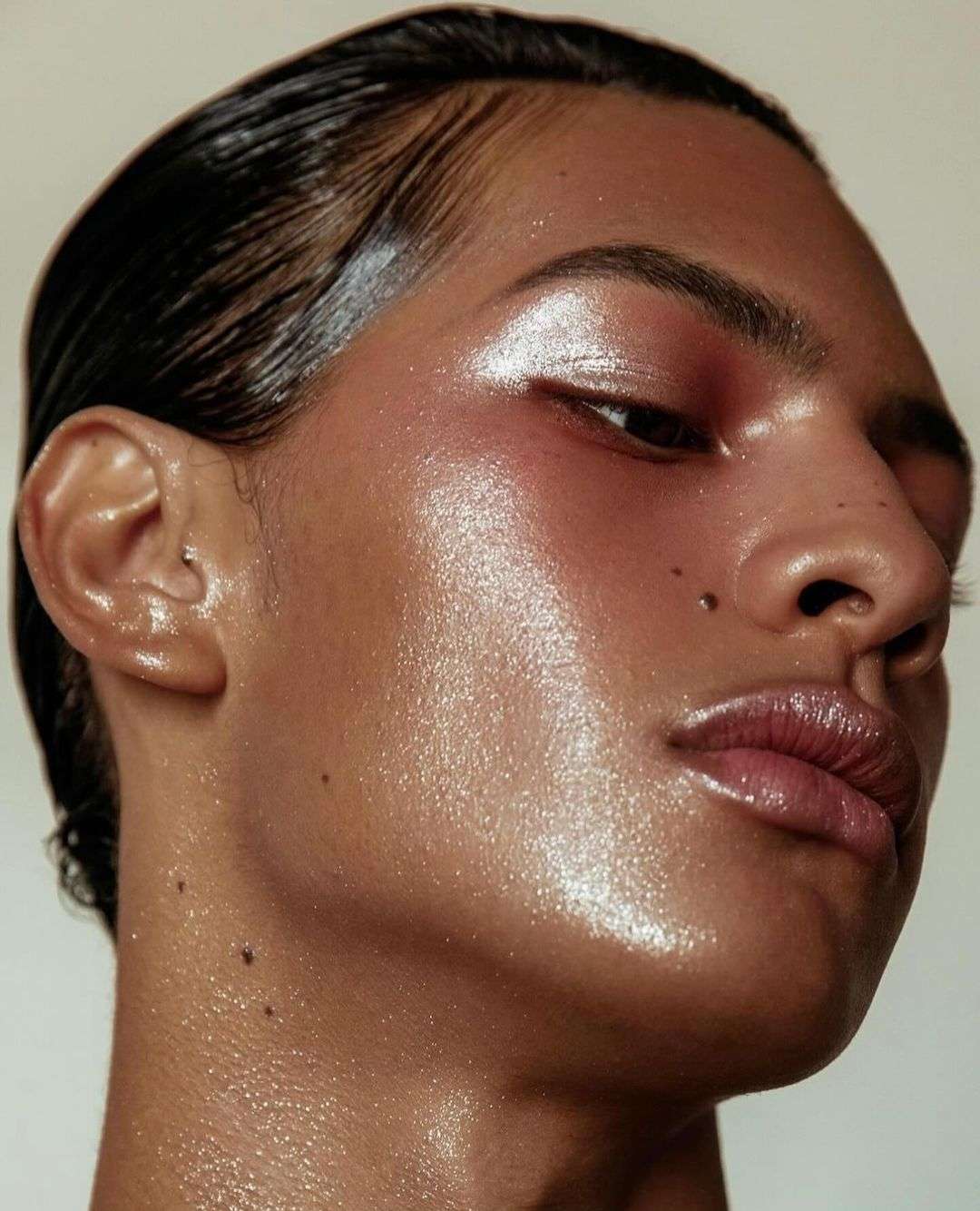 Close-up portrait of a person with wet slicked-back hair and a dewy skin finish, with radiant makeup highlighting their features.