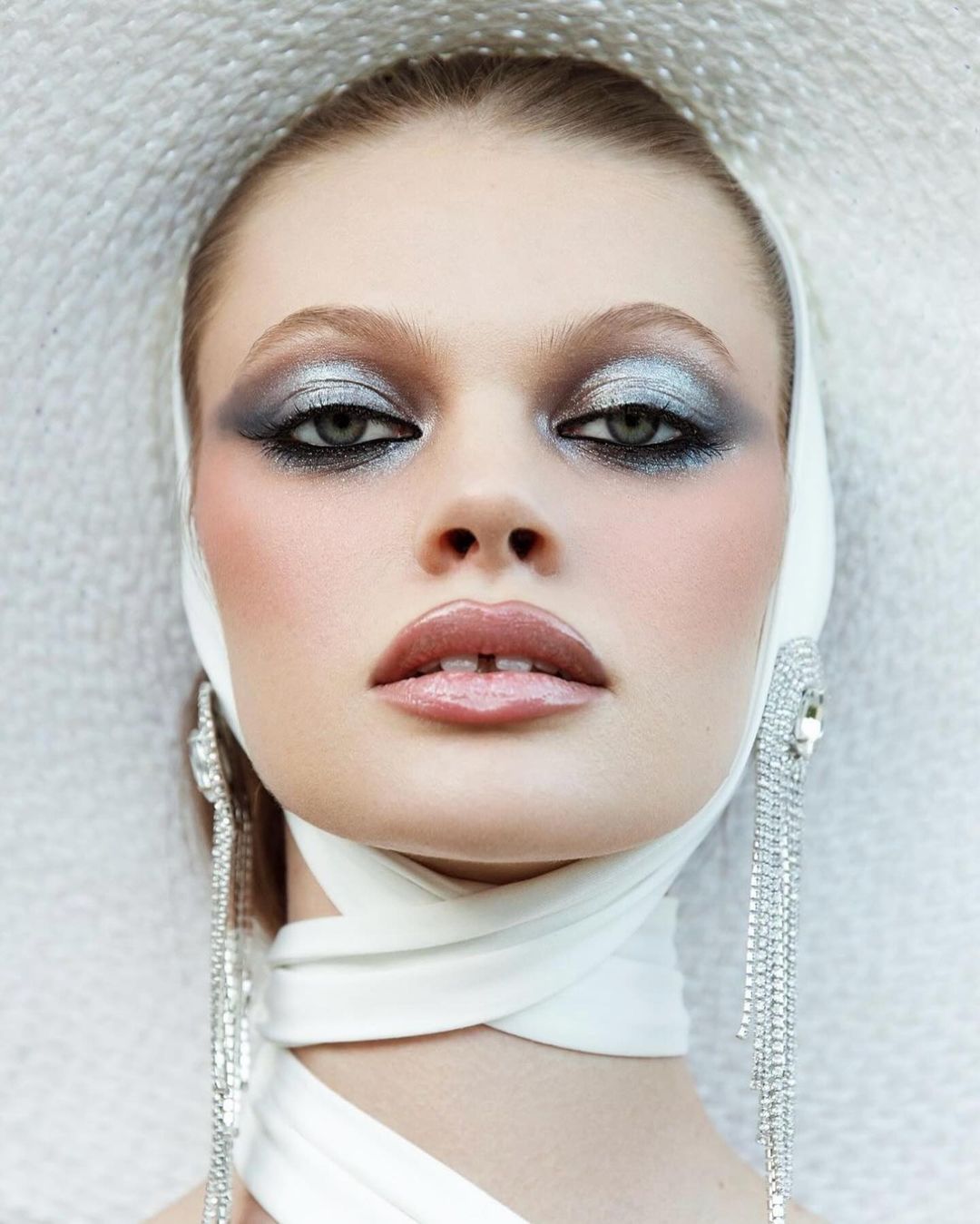 Close-up portrait of a woman with dramatic silver and black eyeshadow, wrapped in a white headscarf and wearing long, sparkling earrings.