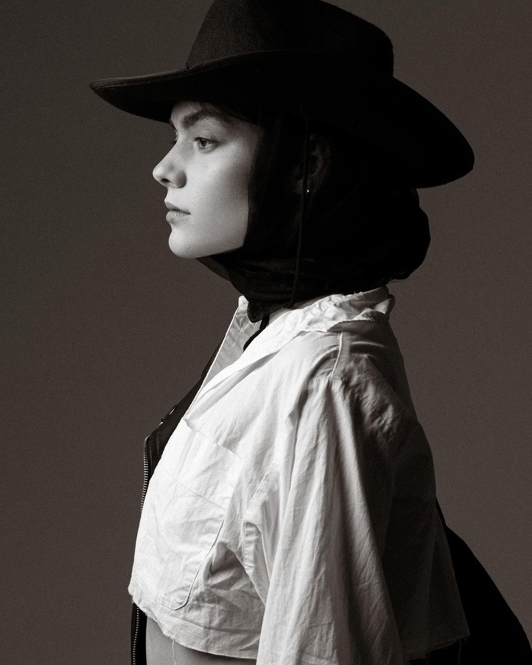 Monochrome portrait of a young woman in a white shirt and black cowboy hat, her face in profile against a gray background.