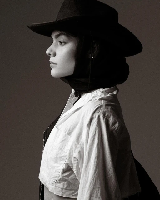 Monochrome portrait of a young woman in a white shirt and black cowboy hat, her face in profile against a gray background.