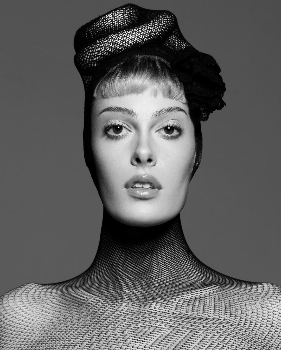 Monochrome close-up studio portrait of a woman with pale skin wearing a black net head covering and shoulder covering.