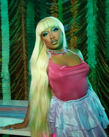 A woman with long pale blonde hair wearing a pink top and light blue ruffled skirt posing against a green backdrop.