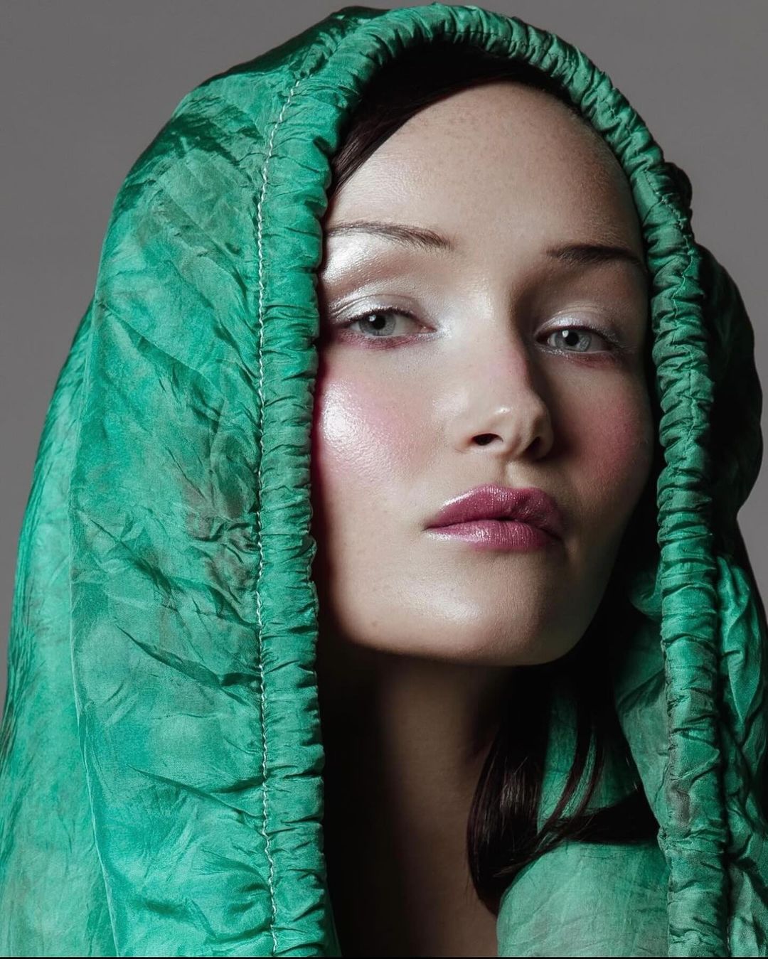 Close-up portrait of an adult female with fair skin, green eyes, and pink lips, wearing a draped green hood and silver eyeshadow.