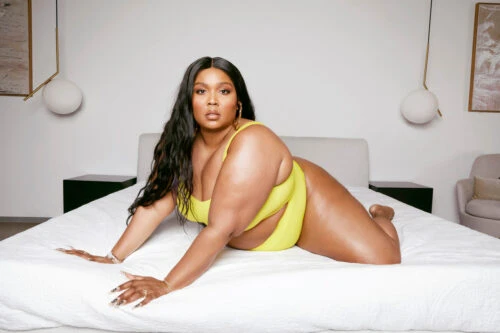 A curvaceous woman with long dark hair poses on a bed in a yellow swimsuit, looking at the camera in a bright, minimalist bedroom.