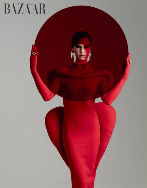 Elegant woman in a red haute-couture gown with matching gloves, makeup, and round headdress, posing against a soft grey backdrop.