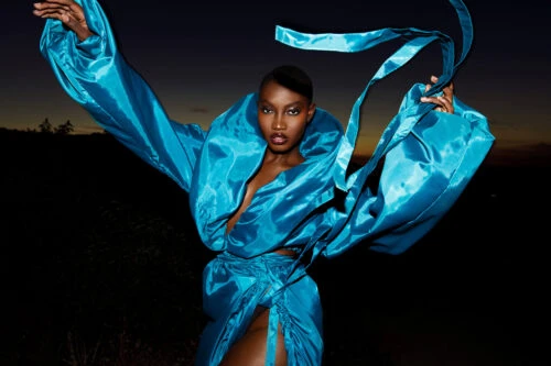 Fashion shot of an adult Black woman in a flowing turquoise dress with a dramatic collar against a dark outdoor background at dusk.