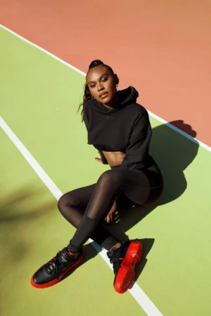 A woman in a black hoodie and leggings poses sitting on a colorful geometric background, showcasing fashion and athletic style with red and black sneakers.
