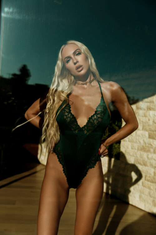 A fair-skinned woman with blonde hair wearing a dark green lace bodysuit poses with hands on hips. The photo shows natural light.