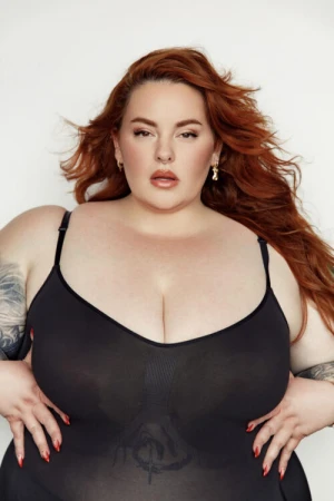 Headshot of a plus-size woman with red hair posing in a black slip dress against a white background in a studio with soft lighting.