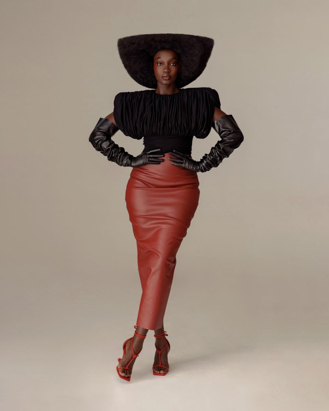 Model wearing a structured black top, red leather midi skirt, oversized hat, and long gloves against a neutral studio backdrop.