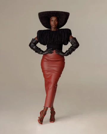 Model wearing a structured black top, red leather midi skirt, oversized hat, and long gloves against a neutral studio backdrop.