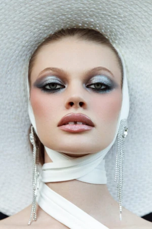 Close-up of a woman wearing dramatic eye makeup, large dangling earrings, and a white ribbon and hat, focusing on beauty and fashion.