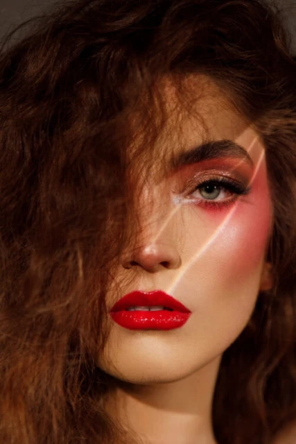 Close-up portrait of a woman with curly brown hair, red lipstick and eye makeup, highlighted by white light streak across her face.