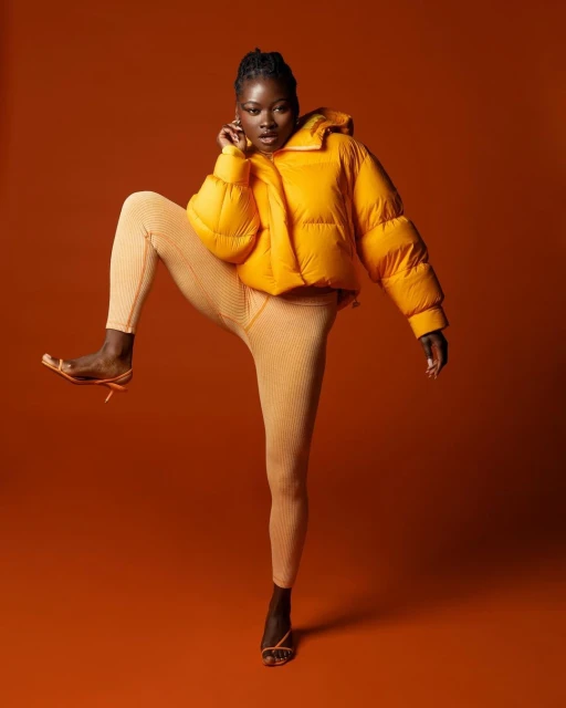 A Black female model poses dynamically in a yellow puffer jacket and matching leggings against an orange backdrop in a studio setting.