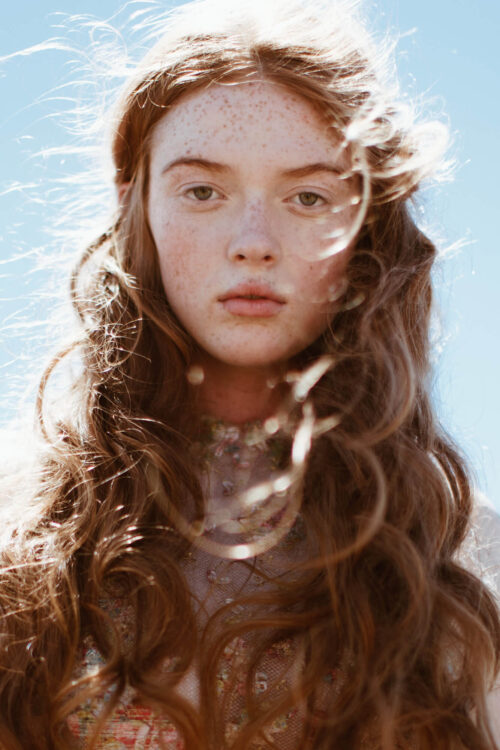 Close-up portrait of a young woman with red hair and freckles against a clear blue sky, emphasizing natural beauty and outdoor aesthetic.