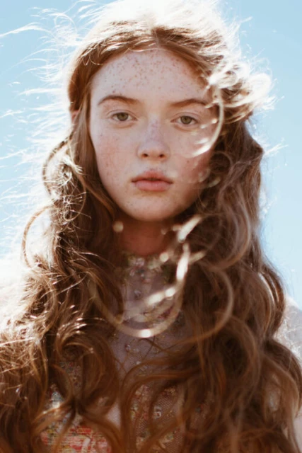 Close-up portrait of a young woman with red hair and freckles against a clear blue sky, emphasizing natural beauty and outdoor aesthetic.